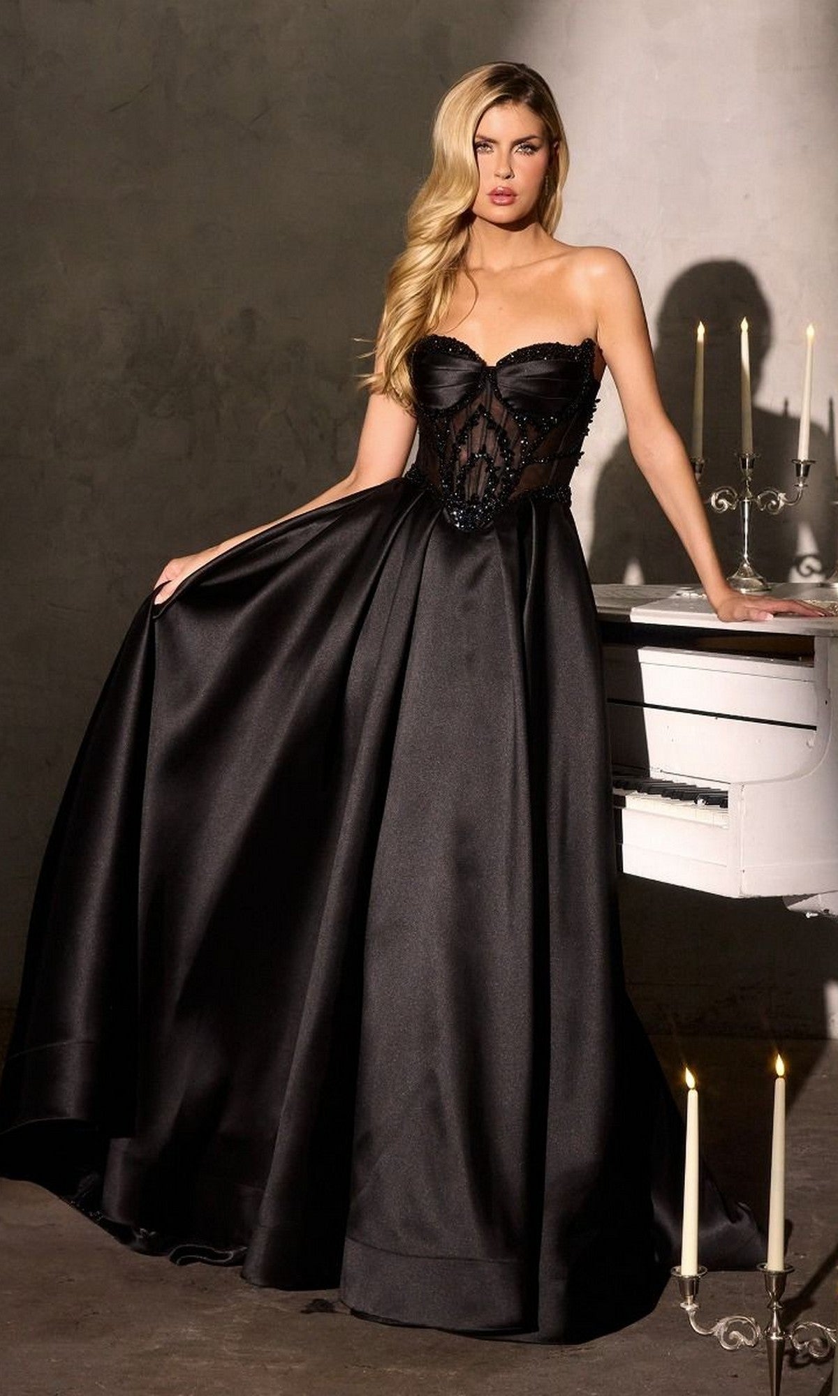 A woman in the black Ladivine CD389 long prom dress, featuring a strapless Mikado design with a sheer bodice and elegant A-line skirt, stands beside a white piano and candles in a dimly lit room.